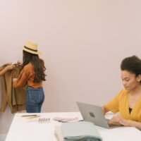 Three women engaged in creative fashion work on laptops and clothing design indoors.