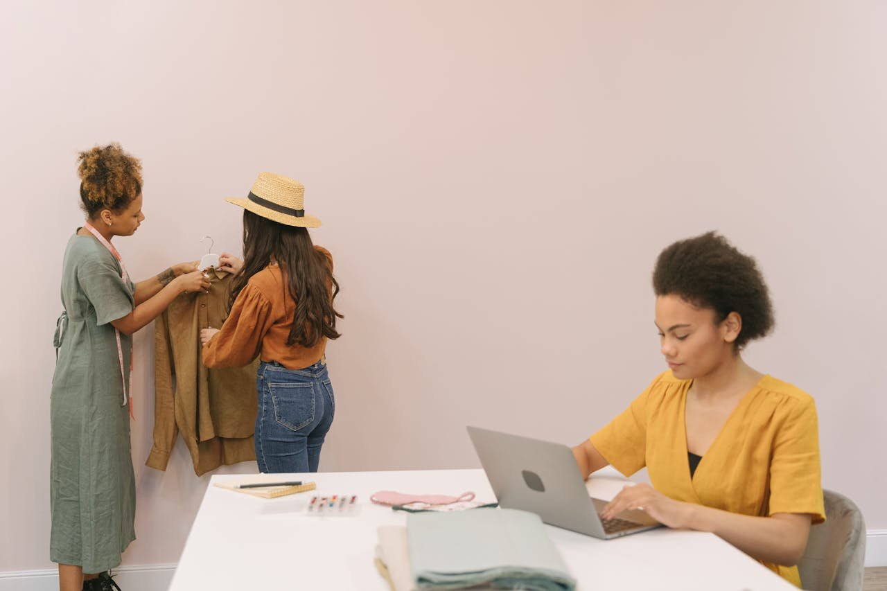 Three women engaged in creative fashion work on laptops and clothing design indoors.